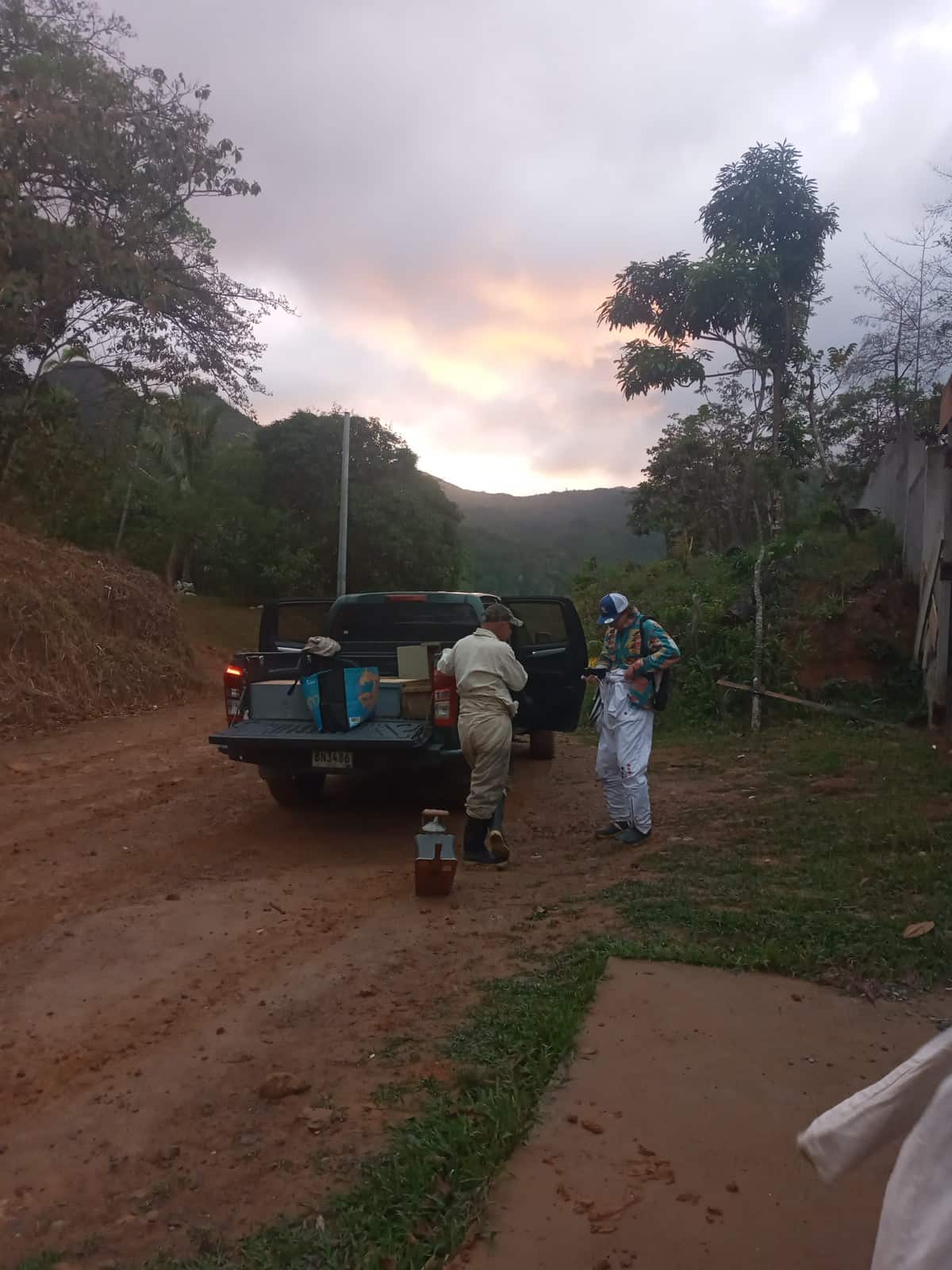 La camioneta parada en el camino de tierra antes de que salga el sol, con el equipo asomando de la batea y dos personas de espaldas junto a ella; detrás, la cordillera y una franja de cielo naranja