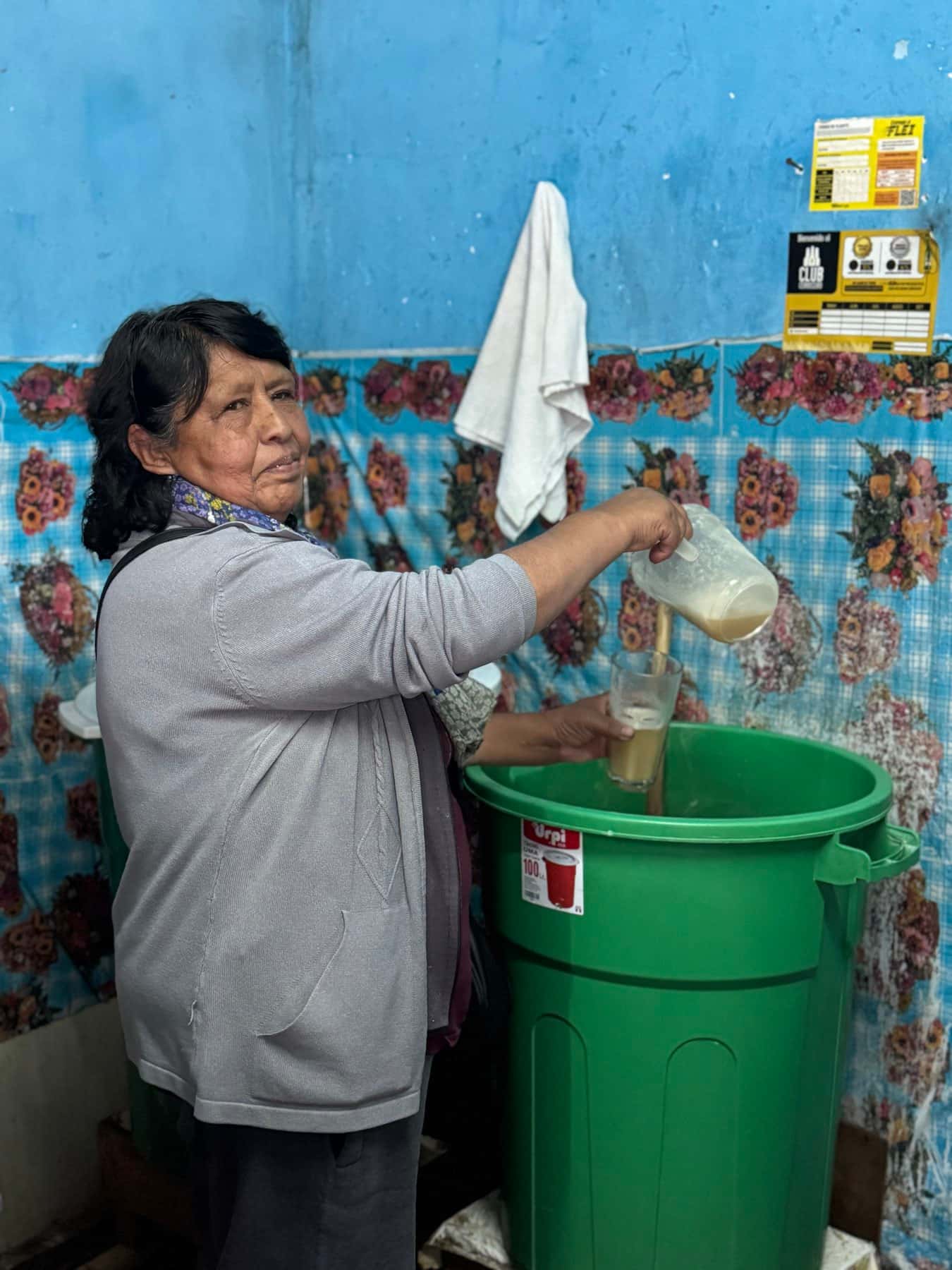 Una mujer sirviendo chicha de una jarra a un vaso, de pie junto a un barril verde, en una pared azul con hule floreado