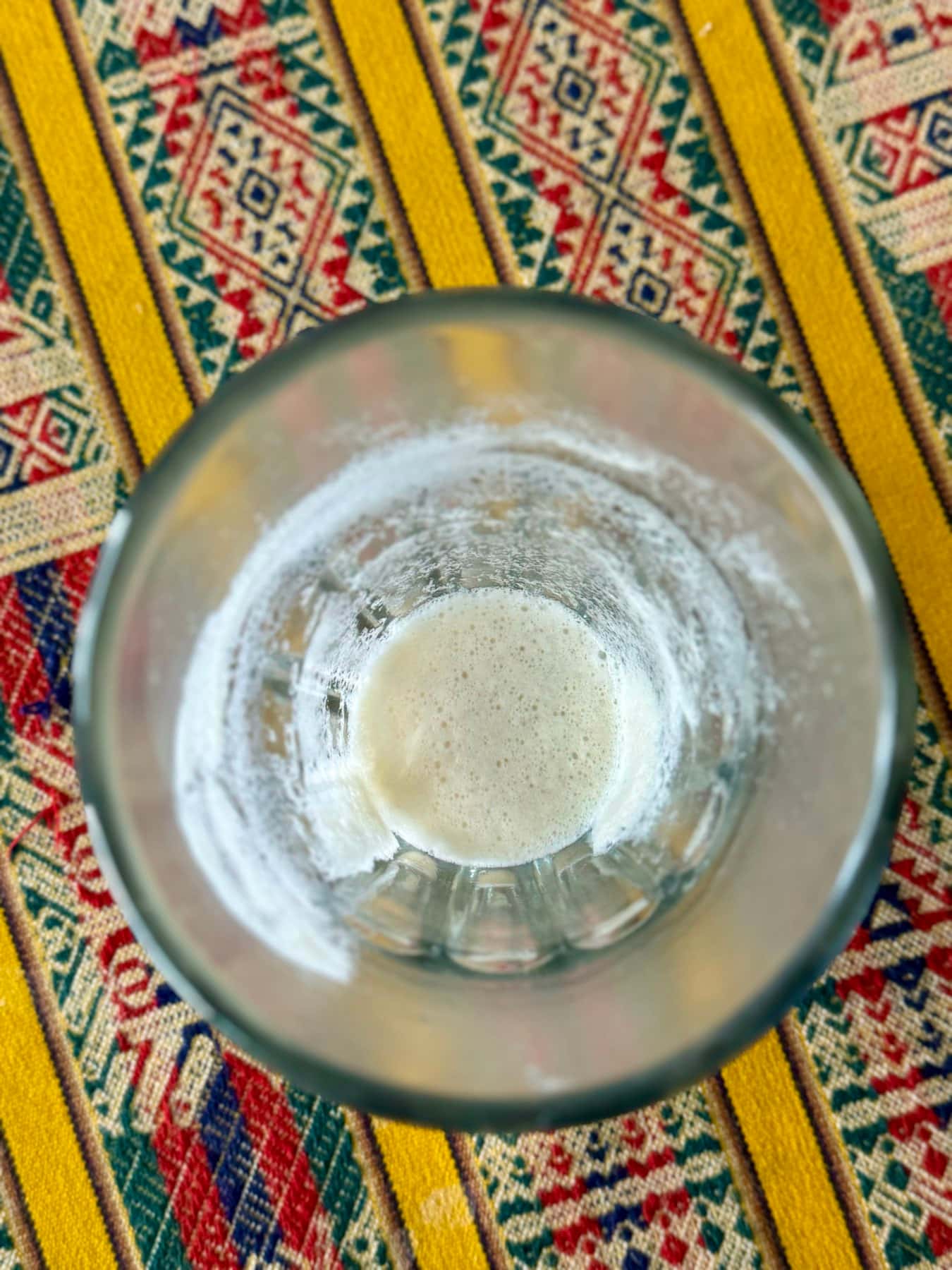 Un vaso casi vacío visto desde arriba, con el poso y la espuma en el fondo, sobre un tejido andino de rayas de colores
