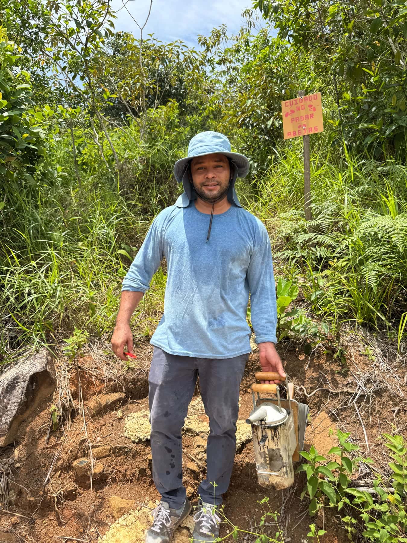 Chayanne López en un sendero de monte, con sombrero de ala y camisa azul, junto a una herramienta y un letrero pintado a mano