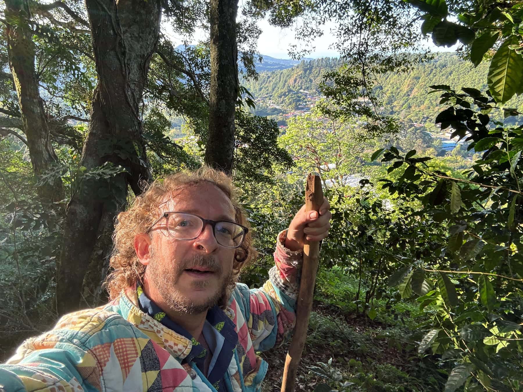 Daniel Giráldez se hace una foto en el bosque, con gafas y una chaqueta de retazos de colores, apoyado en un bastón de madera; al fondo, montañas y un valle
