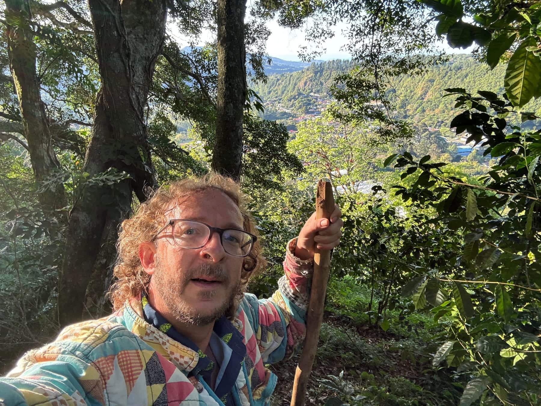 Daniel Giráldez se hace una foto en el bosque, con gafas y una chaqueta de retazos de colores, apoyado en un bastón de madera; al fondo, montañas y un valle