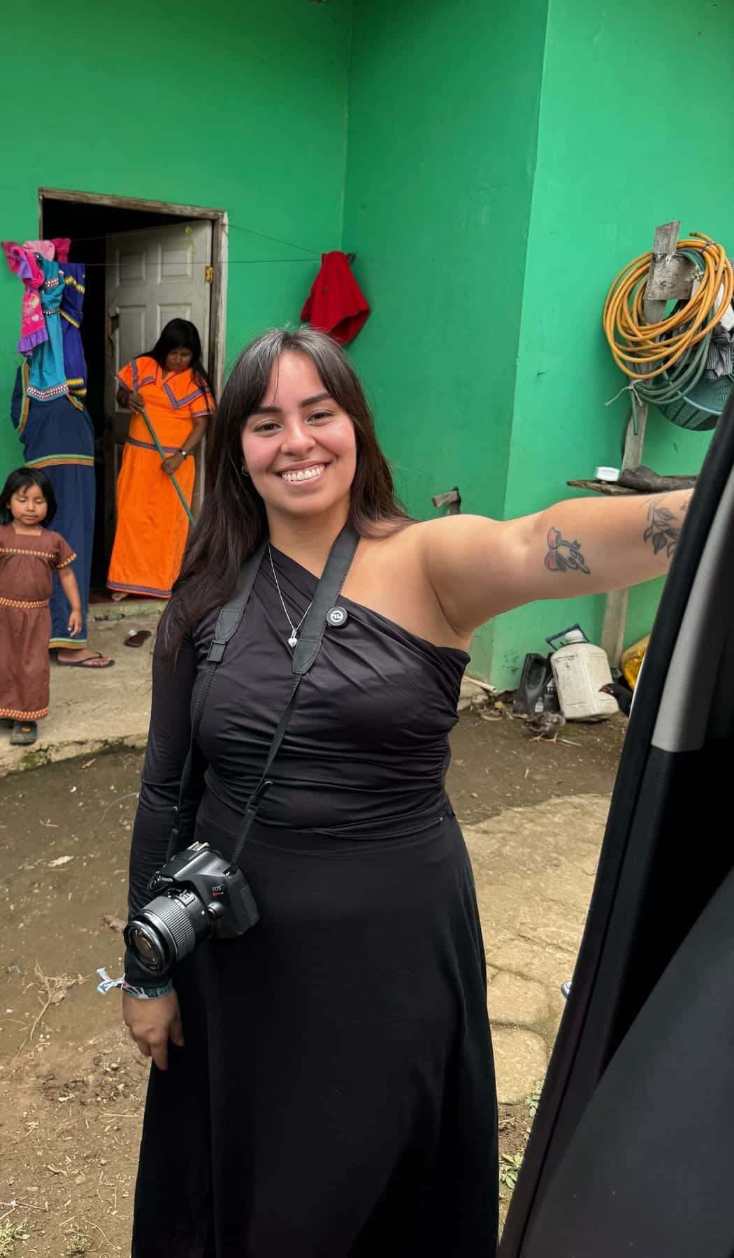 Nathy Ruiz sonríe con el brazo extendido y una cámara colgada del hombro, frente a una casa verde; al fondo, dos personas junto a la puerta