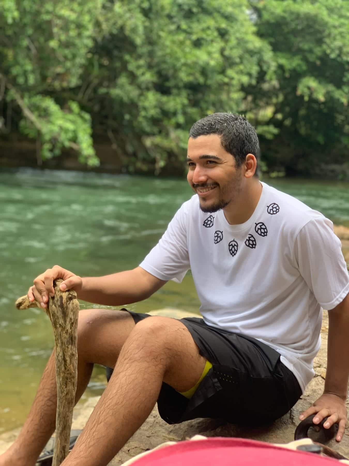 Oliver Chez sentado junto a un río, con una camiseta blanca y un palo de madera en la mano