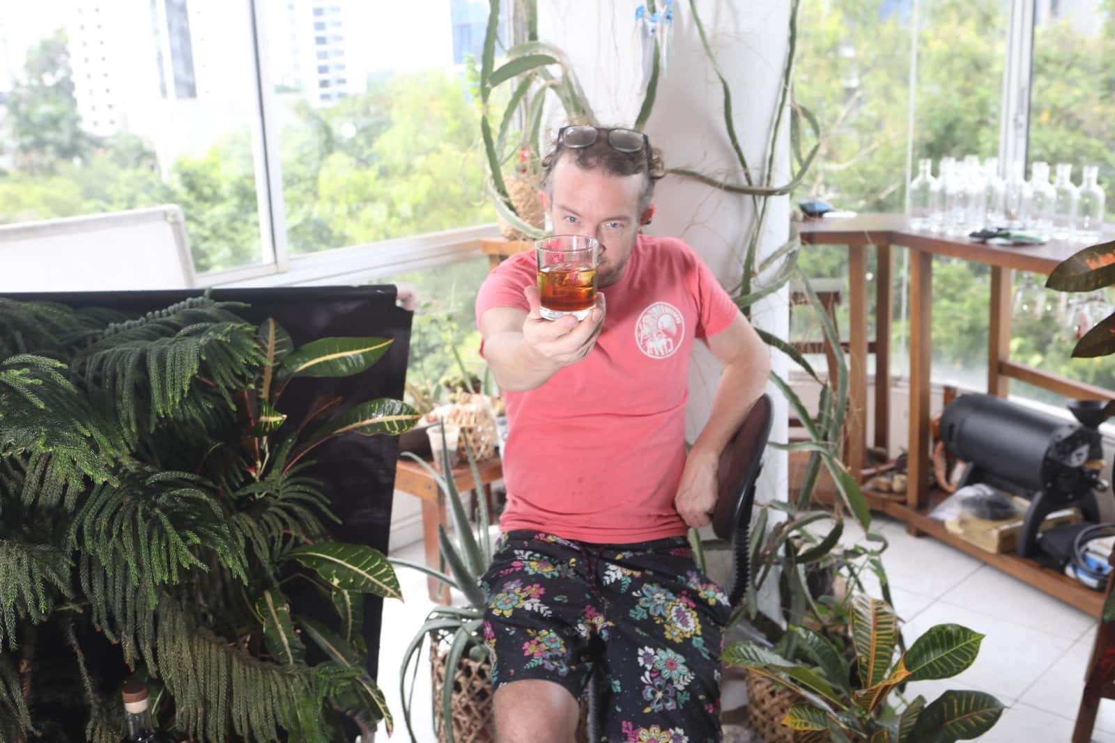 Daniel sentado en una terraza entre plantas, ofreciendo hacia la cámara un vaso servido