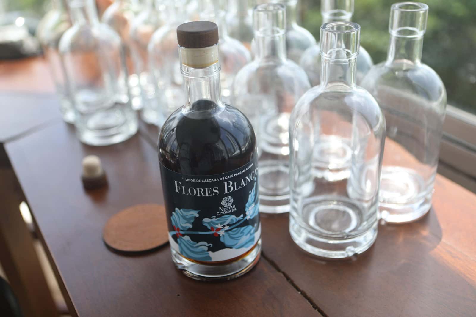 La botella etiquetada sobre una mesa de madera, entre botellas vacías y cristalería, junto a la ventana