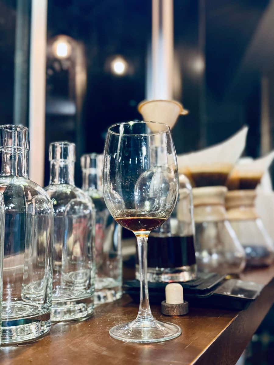 Una copa de cata con un licor ámbar, entre botellas vacías y un embudo, sobre una mesa de madera