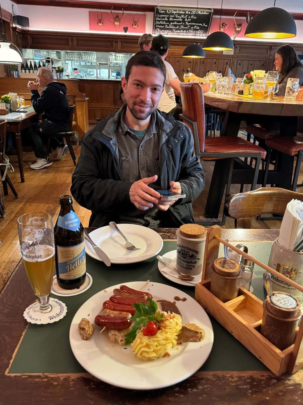 Un hombre sentado a una mesa de madera con una cerveza Weihenstephaner Helles, un plato de salchichas con chucrut y puré, y al fondo la barra del local y una pizarra