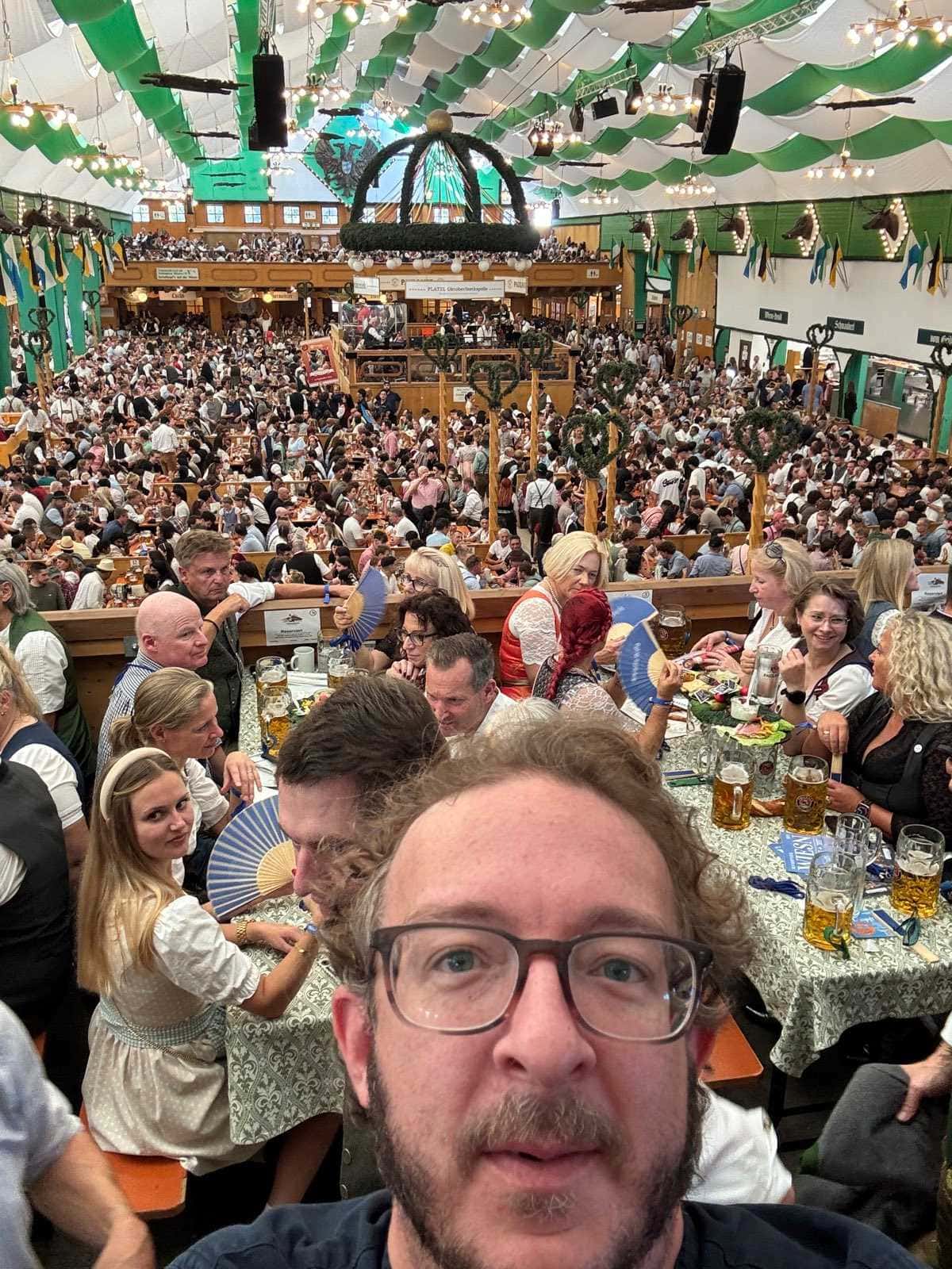 Interior de una carpa del Oktoberfest vista desde arriba: cientos de personas en mesas corridas bajo un techo de tela verde y blanca, con jarras de litro sobre los manteles