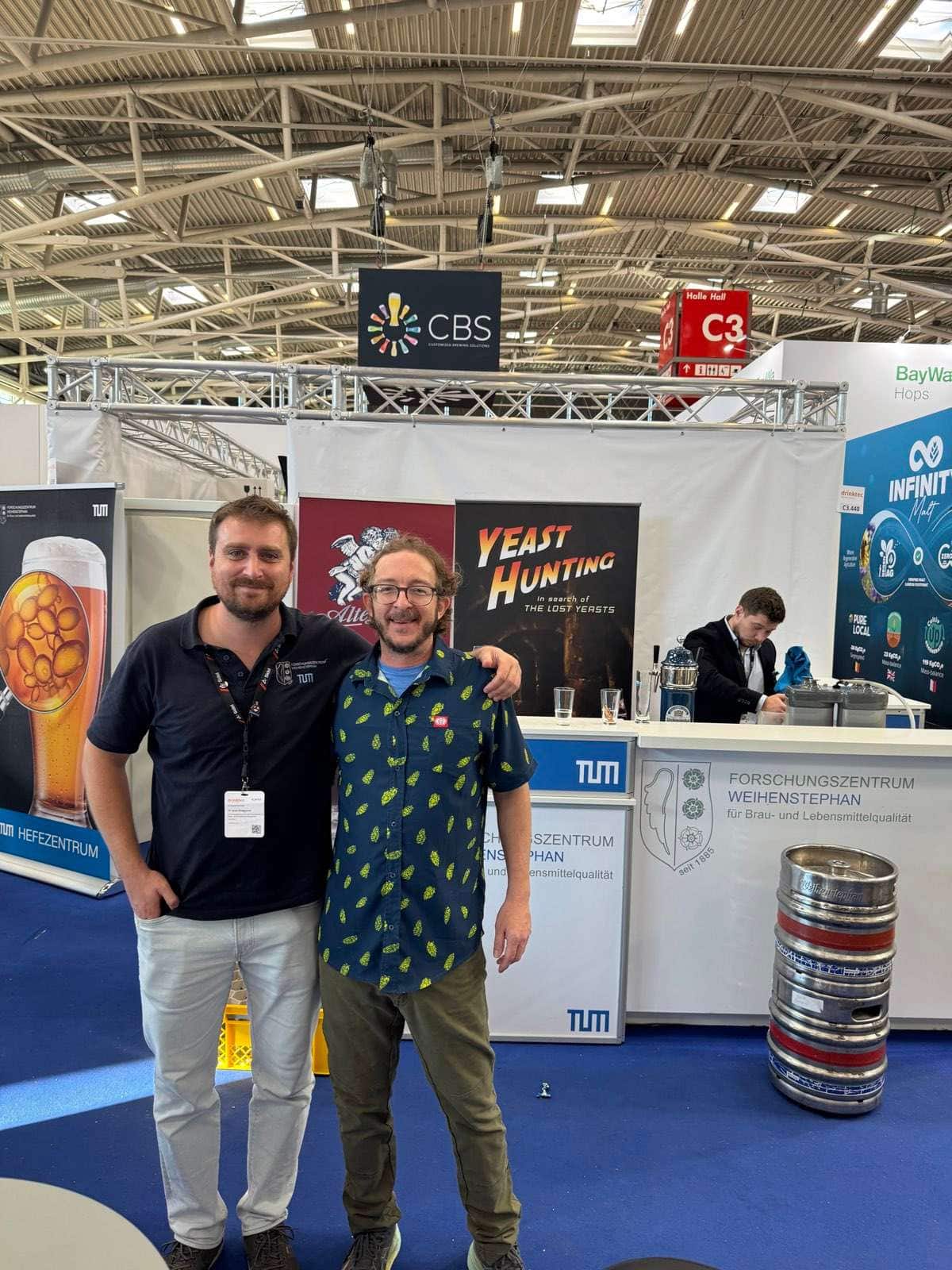 Dos hombres posando en el pasillo de una feria, delante de un estand con un cartel que dice «Yeast Hunting, in search of the lost yeasts» y los rótulos del centro de investigación de Weihenstephan y del Hefezentrum de la TUM