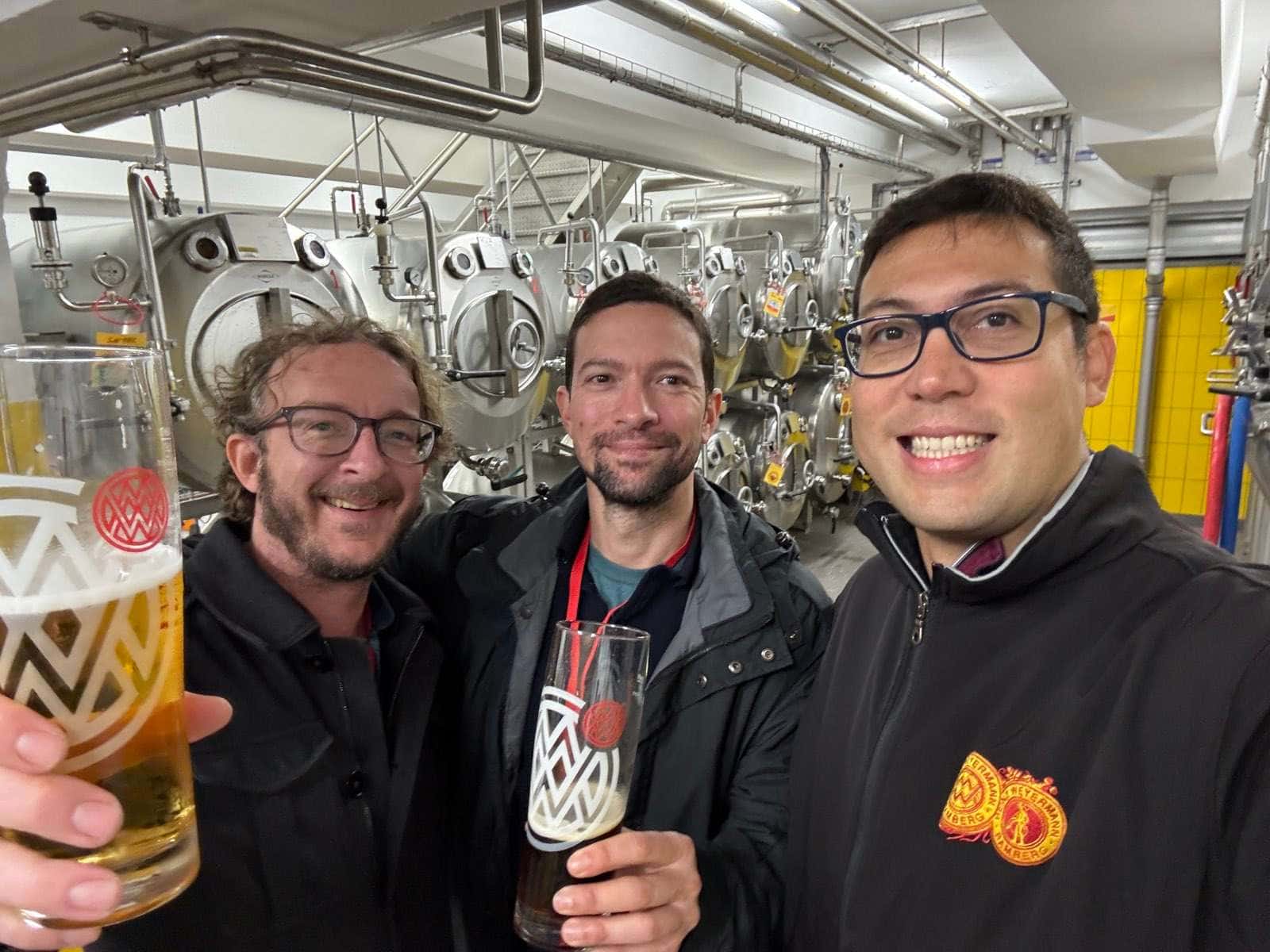 Tres hombres en la bodega de una cervecería, brindando con copas de Weyermann; detrás, una hilera de tanques horizontales de acero