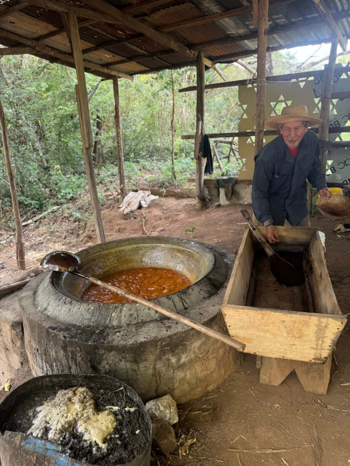 Plano general del trapiche: la paila humeante con el cucharón cruzado encima, la canoa de madera esperando a la derecha, y en primer término un cuenco con la raspadura ya cuajada