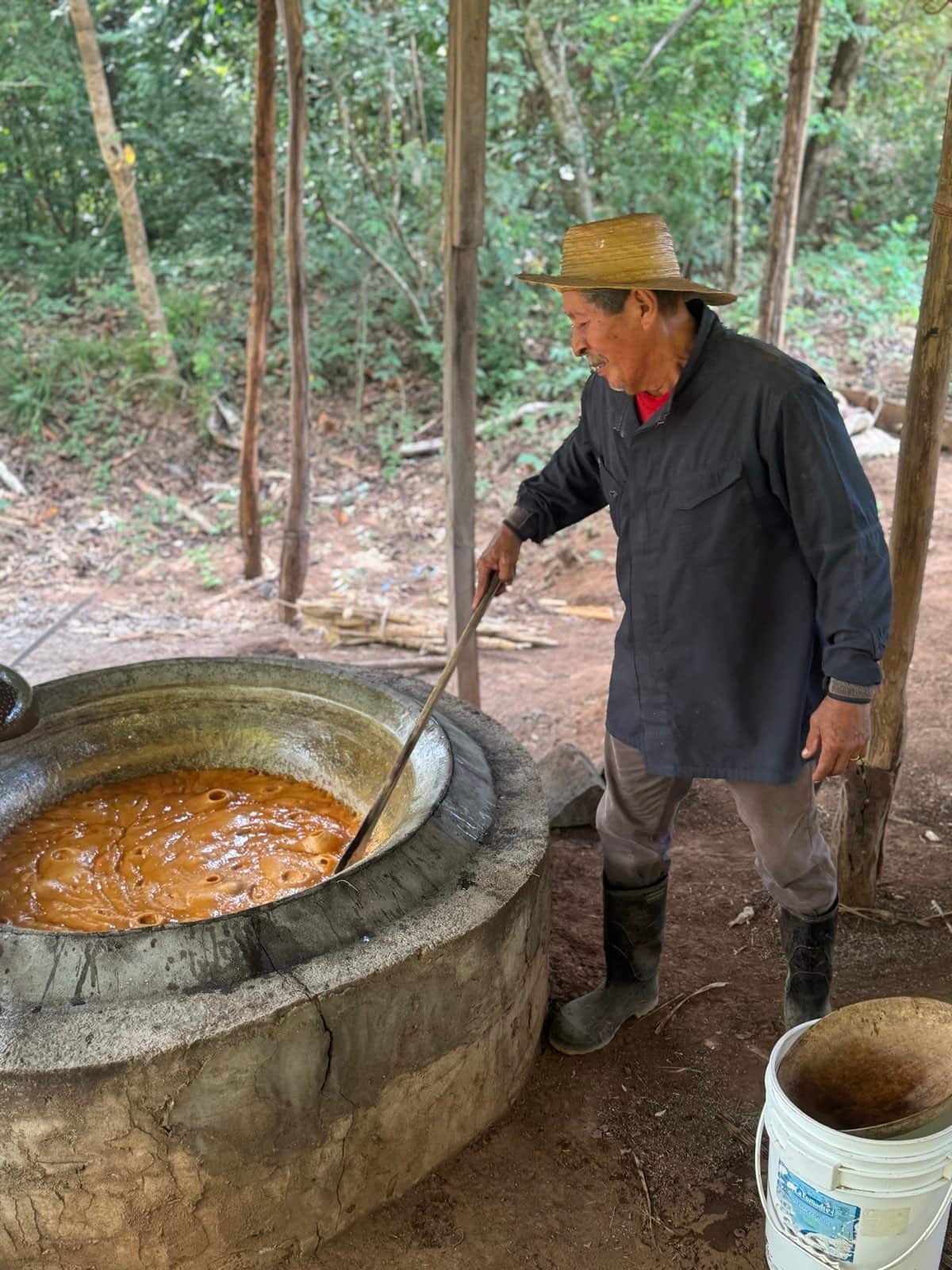 Un hombre con sombrero de paja removiendo con una paleta larga el guarapo hirviendo en una paila de metal, montada sobre un fogón de barro bajo una enramada de madera