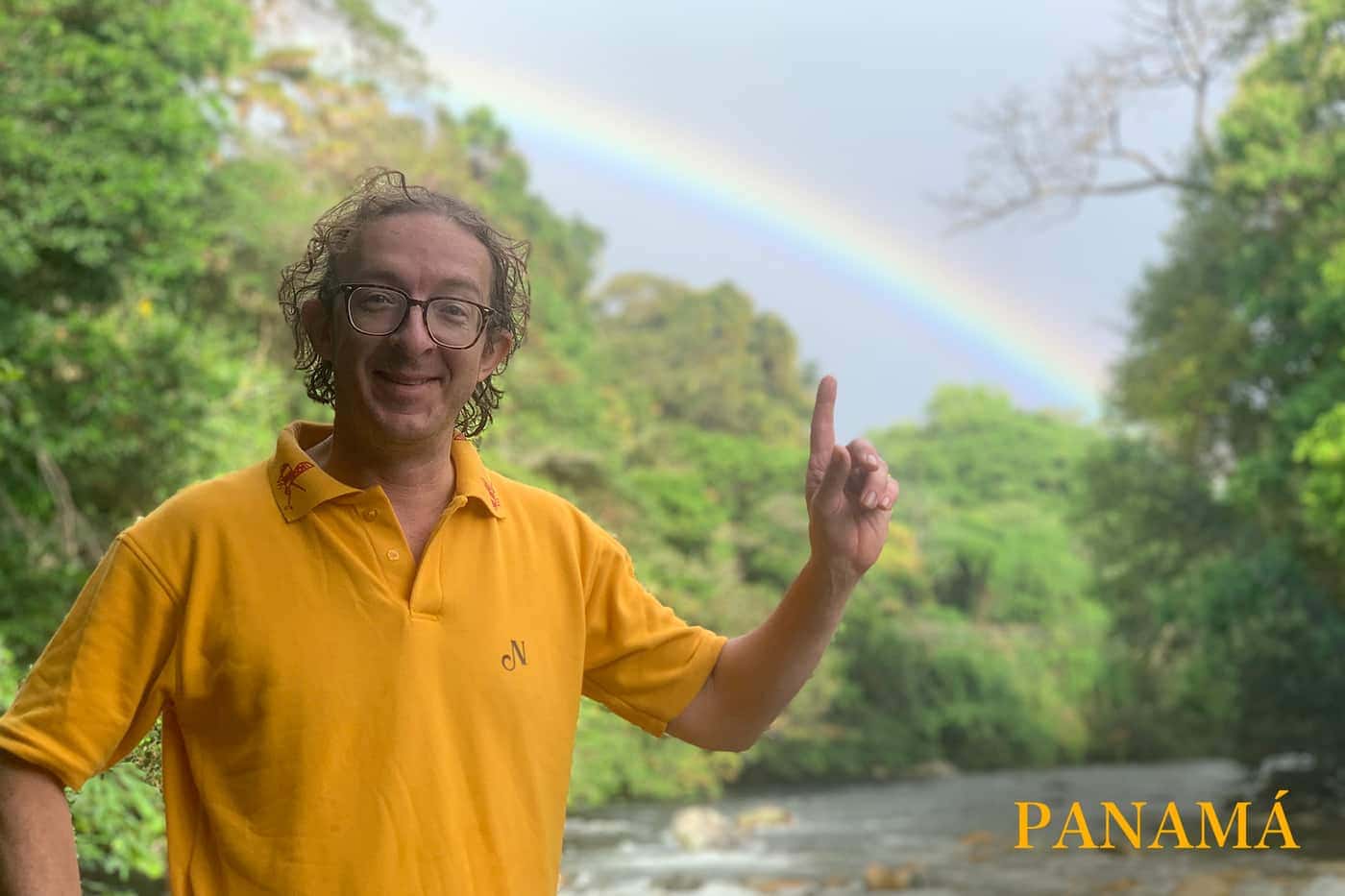 Un hombre de pelo rizado y gafas, con polo amarillo, señalando hacia un arcoíris sobre la vegetación de un río