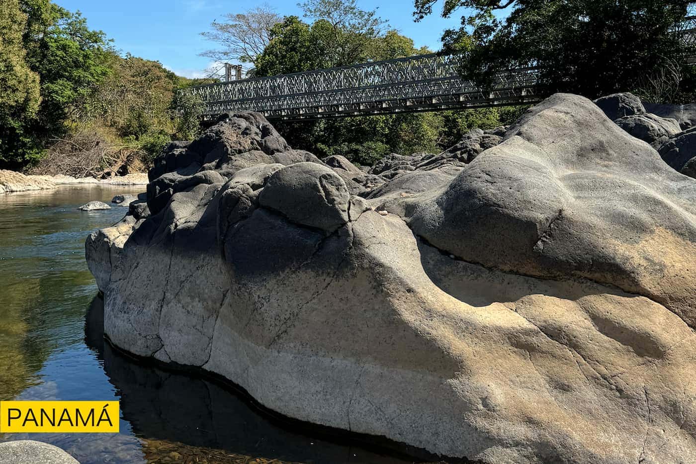 Rocas lisas junto a un río y, al fondo, un puente colgante entre la vegetación